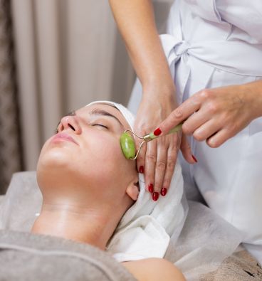 An Ayurvedic therapist preparing herbal medicines while a woman relaxes, using a face roller during a rejuvenating facial massage at Jivika Wellness Spa